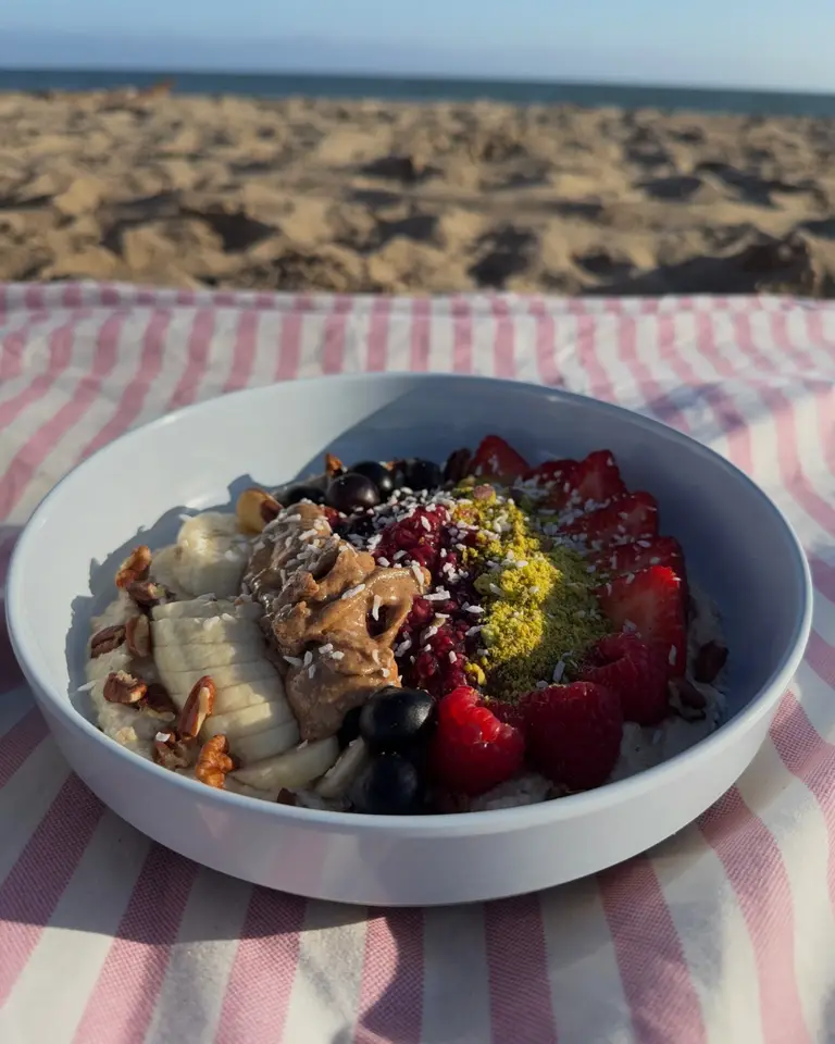 Sante Organics signature bowl with raspberry-chia jam, strawberries, raspberries, banana, pecans and crushed pistachio on a pink striped towel at the beach